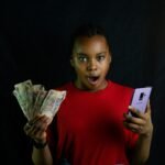 uma jovem negra com notas de dinheiro numa mão e o celular na outra