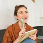 retrato-de-jovem-sorridente-com-pensamentos-criativos-olhando-para-cima-com-rosto-inspirado-segurando-caneta e caderno